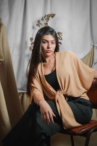A vertical shot of a sophisticated young Latina woman seated in a studio, modeling a tan knotted kimono top over black against a moody fabric backdrop