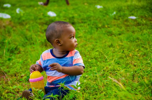 A closeup of cute little Black boy sitting on the grass in the park and looking away