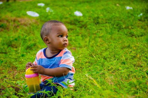 A closeup of cute little Black boy sitting on the grass in the park and looking away