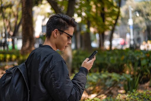 Young teenage boy holding cell phone in his hand and browsing the internet in the park with blurry background.