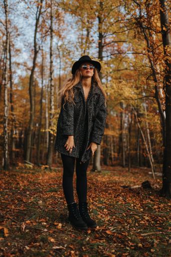 A vertical shot of a blonde sexy female model with black coat and hat posing in an autumnal forest