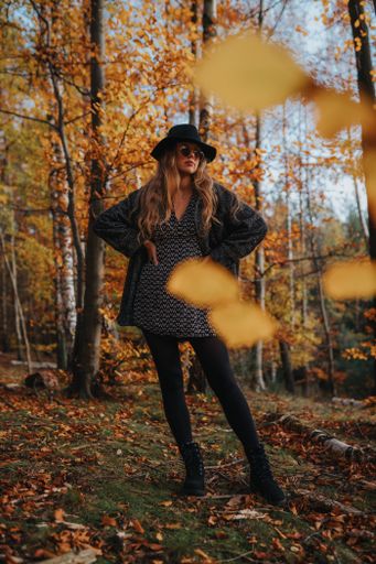 A blonde female model with black coat and hat posing in a forest full of yellow autumnal leave