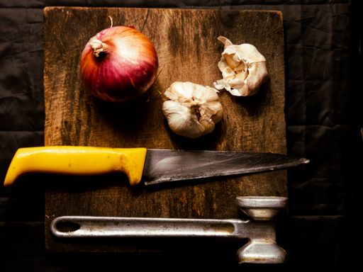A top view of an onion and garlic bulbs on a cutting board with a knife and a hammer on it