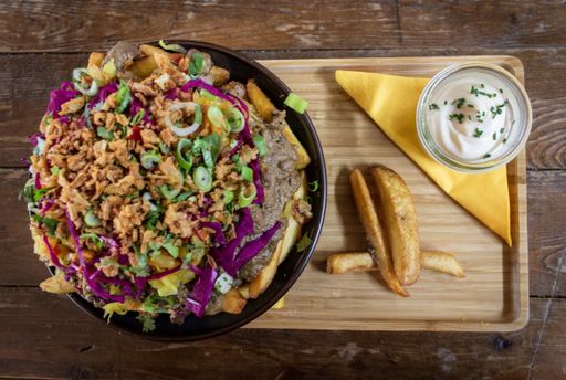 An overhead shot of a dish with potatoes, meat and sliced vegetables in a bowl near a sauce on a wooden tray