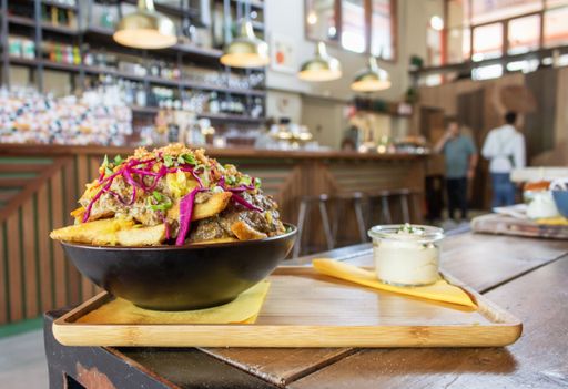 A closeup of a dish with potatoes, meat and sliced vegetables in a bow near the sauce on a wooden tray with a blurred background