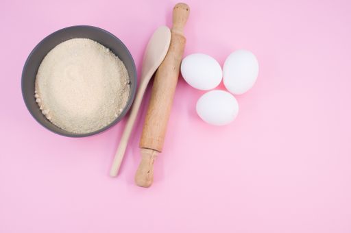 wooden rolling pin white eggs and dark gray bowl with wholegrain flour on pink background with copy space
