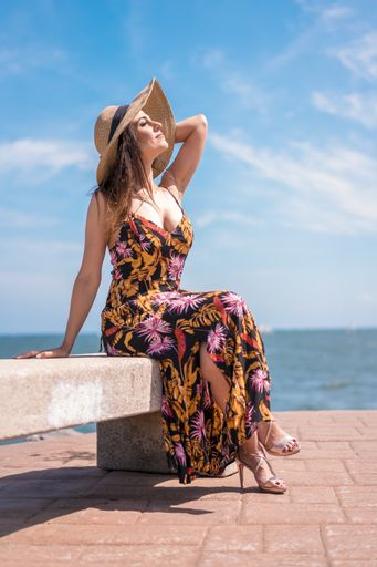 A vertical shot of a female in a floral sundress and a hat sitting by the sea captured in Spain