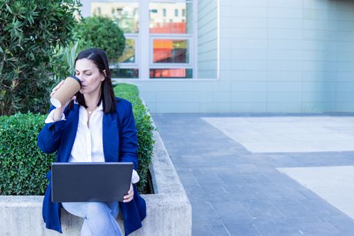 Beautiful Caucasian businesswoman using a laptop computer and drinking coffee sitting outside an office building, copy space
