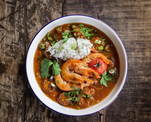 An overhead shot of soup with shrimps and vegetables leaves in a white bowl with a wooden surface in the background