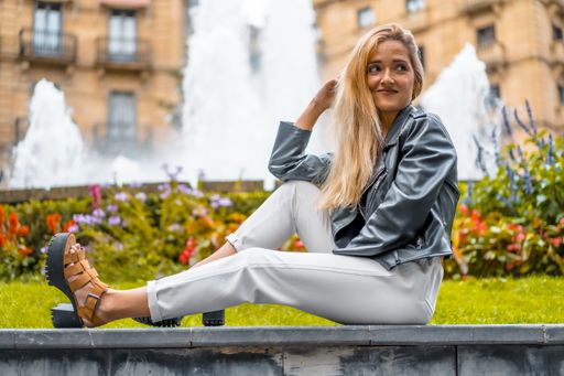 A female in white pants and leather jacket sitting near the flowers and fountain in San Sebastian, Spain