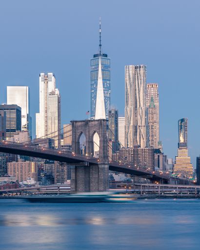 A vertical distant shot of the Brooklyn bridge on the body of water near skyscrapers in New York