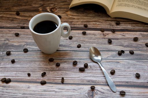 A cup of coffee, a spoon, and a book on a wooden surface with scattered coffee beans