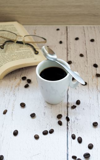 A vertical shot of a cup of coffee, a spoon, a book, and glasses on a wooden surface with scattered coffee beans