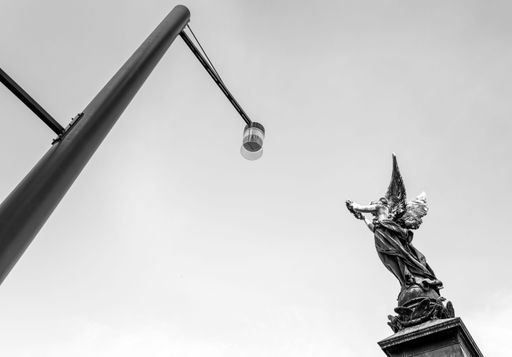 A grayscale shot of a statue of a winged angel and lamppost against a bright sky in Freiburg im Breisgau, Germany during daytime