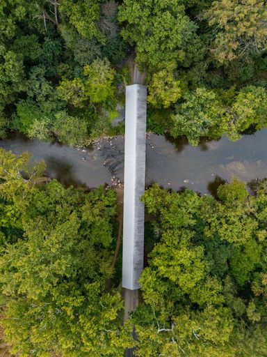 Aerial view of a covered bridge in a lush green forest over a flowing river, showcasing nature's beauty and architectural charm.