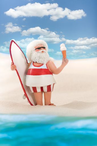 A relaxed Santa about to go surfing, he is standing on the shore of a sandy beach eating ice creambefore going in the water.