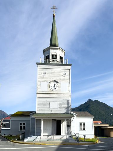 Historic church with green spire and clock tower set against blue sky and mountain backdrop in Sitka Alaska