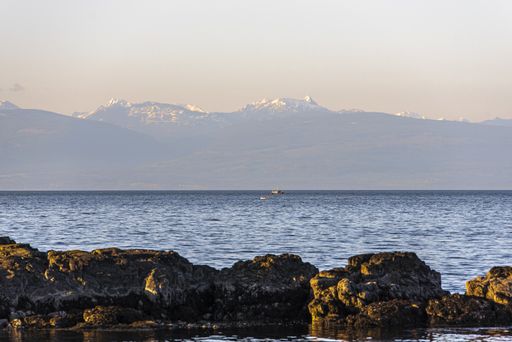 A wide shot of rock formations near the body of water surrounded by mountains in the distance under a pink sky