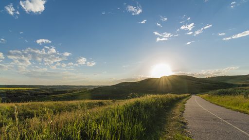 A  highway road near grass fields surrounded by a mountain the distance under a sunny blue sky