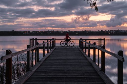 A bicyclist standing on a wooden dock on the water under a cloudy sky during the sunset in the evening