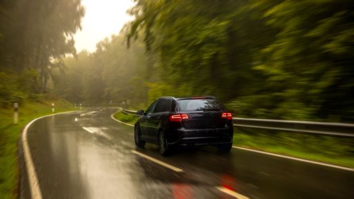 A sleek car drives on a wet, winding road through a lush, green forest, creating a sense of motion and adventure.