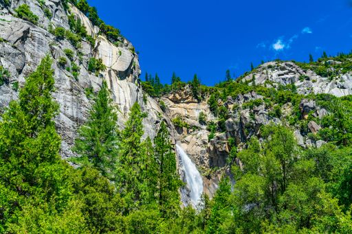 Scenic view of a waterfall cascading down a rocky cliff surrounded by lush greenery and a vibrant blue sky in Yosemite National Park.