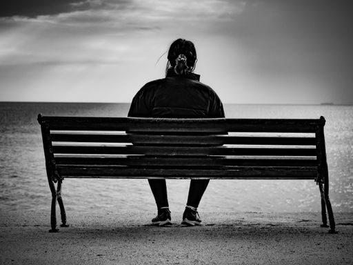 A grayscale shot of a woman sitting on the bench and looking at the sea