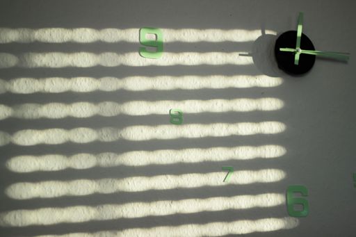 A minimalist wall clock with green numbers and shadow patterns from blinds on a white wall, creating an abstract design.