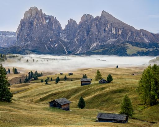 A stunning view of the Dolomites with mist over rolling green hills and scattered wooden huts in Alpe di Siusi, Italy.