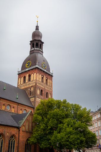 View of Riga Cathedral with its iconic spire and clock tower against a cloudy sky, featuring lush green trees in the foreground.