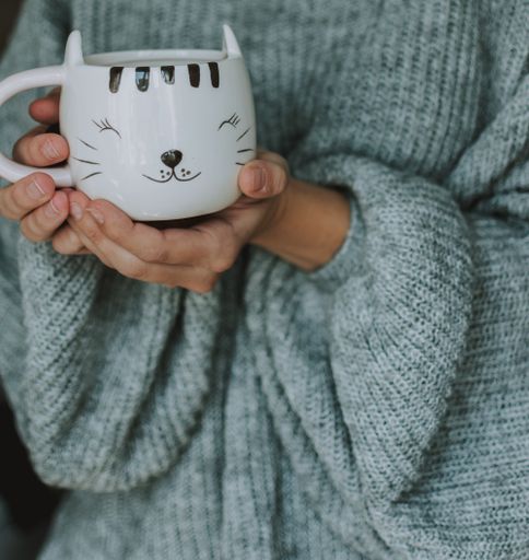 A closeup shot of a female wearing blue sweater holding a cute white mug with the face of a happy kitten