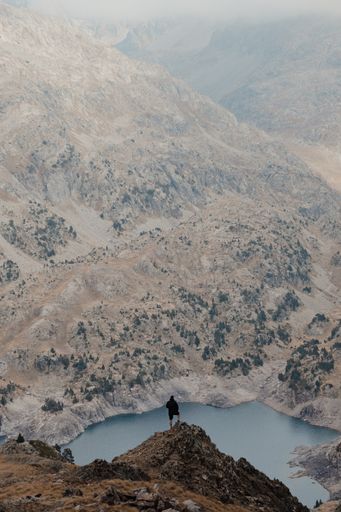 A vertical shot of a hiker standing on a cliff and observing a beautiful mountainous landscape with a lake