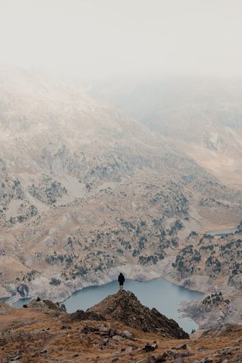 A vertical shot of a hiker standing on a cliff and observing a beautiful mountainous landscape with a lake