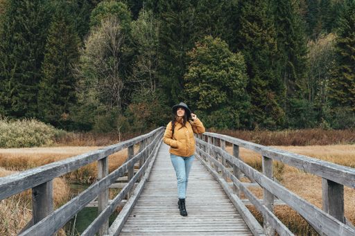 Young woman wearing yellow jacket and fedora hat, standing on wooden path across grassland with creek at Zelenci nature reserve near Kranjska gora.