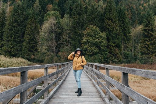 Young woman wearing yellow jacket and fedora hat, standing on wooden path across grassland with creek at Zelenci nature reserve near Kranjska gora.