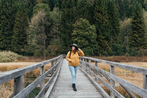 Young woman wearing yellow jacket and fedora hat, standing on wooden path across grassland with creek at Zelenci nature reserve near Kranjska gora.