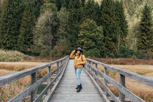 Young woman wearing yellow jacket and fedora hat, standing on wooden path across grassland with creek at Zelenci nature reserve near Kranjska gora.