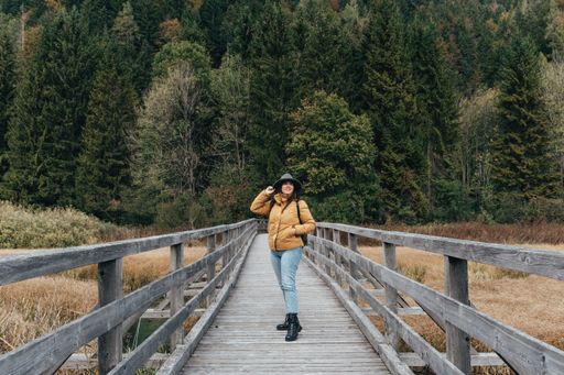 Young woman wearing yellow jacket and fedora hat, standing on wooden path across grassland with creek at Zelenci nature reserve near Kranjska gora.
