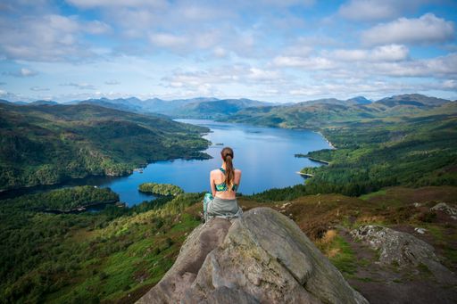 A scenic shot of a woman from her back sitting on a rock on the mountain looking at the calm lake
