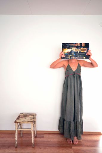 A vertical shot of a female covering her face with an abstract painting, standing against a white wall next to a wooden chair