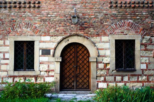 A facade of a brick and stone building with two windows and green plants in the garden