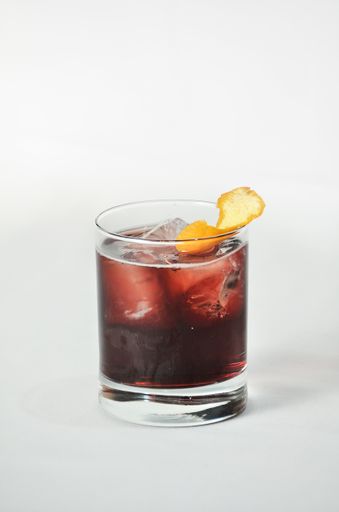 A vertical shot of a dark red liquid in an ice-filled glass with an orange twist on top and white background