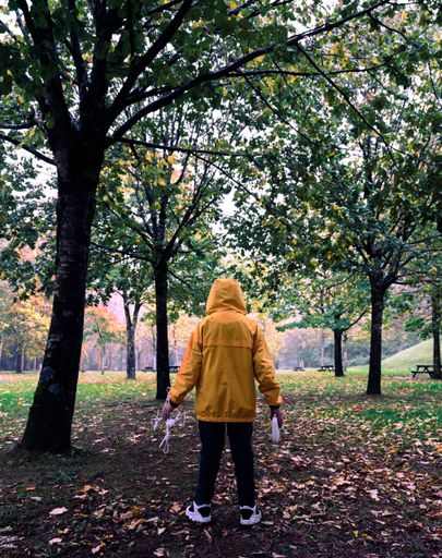 A back view of a little Caucasian boy in a yellow coat holding a drone and remote control in the park