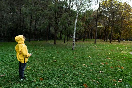 A back view of a little Caucasian boy in a yellow coat flying a drone in the park on a sunny day