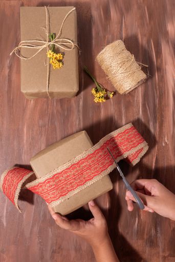 A top view of a person wrapping a Christmas present with jute fabric ribbon with red lace on a table