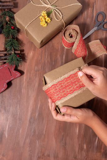 A top view of a person wrapping a Christmas present with jute fabric ribbon with red lace on a table
