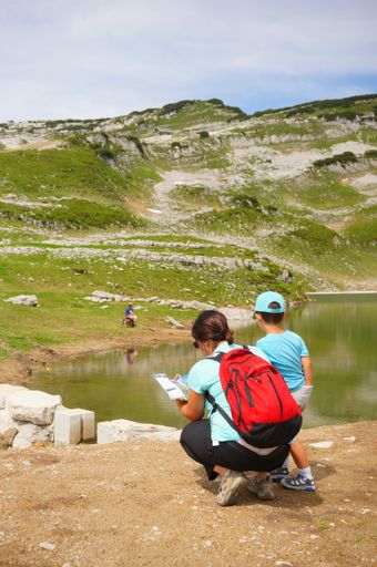 A woman with a backpack and child reading a map in front of a lake on a mountain