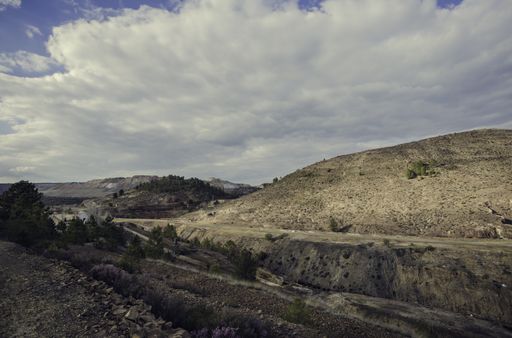 a view of tree-covered rocky hills with dirt road under the cloudy sky on a bright, sunny day