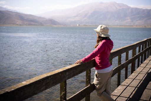 A woman in a sun hat and pink jacket enjoys a lakeside view from a wooden pier, with mountains in the background under a clear sky.