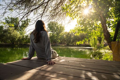A woman in her 20s sits on a wooden deck by a serene pond, surrounded by lush greenery and sunlight filtering through the trees.